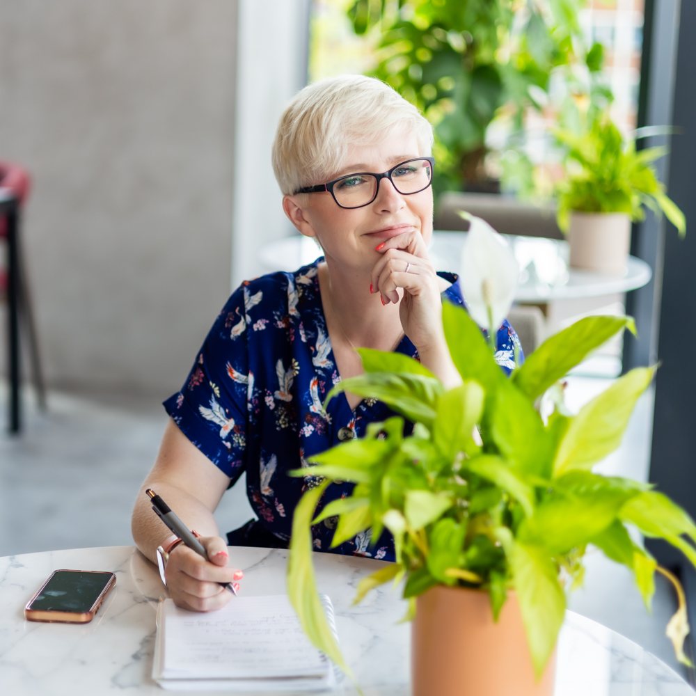 Jenny at a table with a pen and notebook, beside a plant