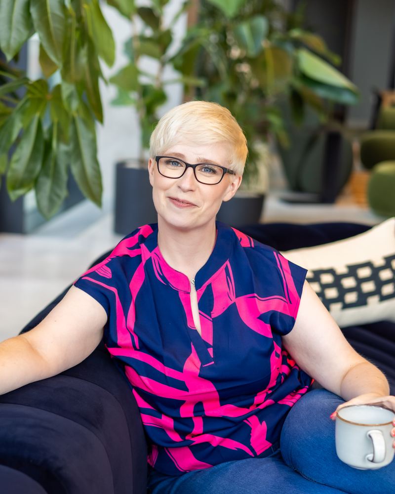 Jenny Lucas sitting on a navy sofa in a navy and pink top, holding a mug and smiling